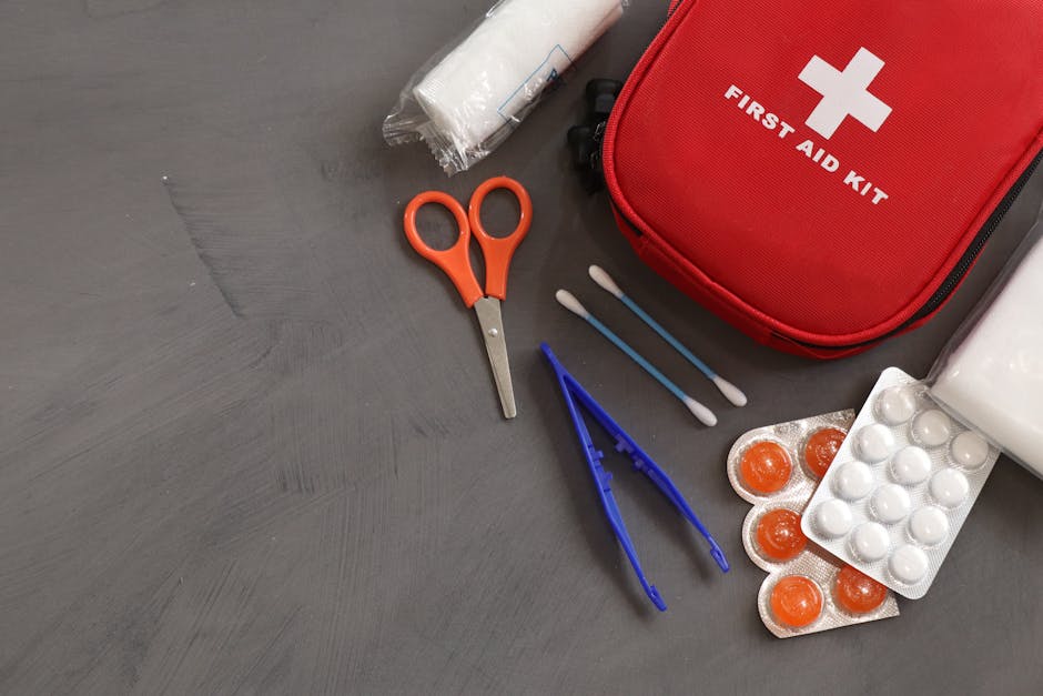 Red first aid kit with a white cross sits among scissors, tweezers, cotton swabs, and blister packs on a gray surface—first-aid supplies ready to use.